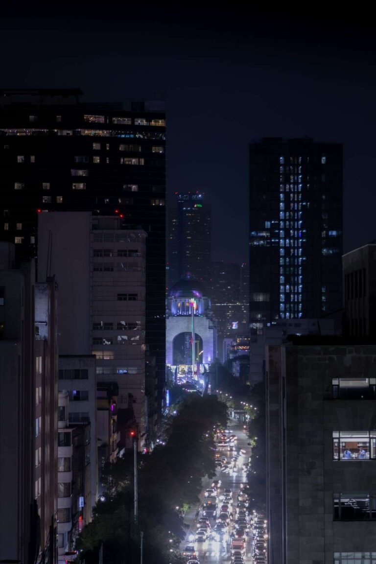 Busy Mexico City avenue at night with traffic trails leading to the lit Monumento a la Revolucion between tall buildings